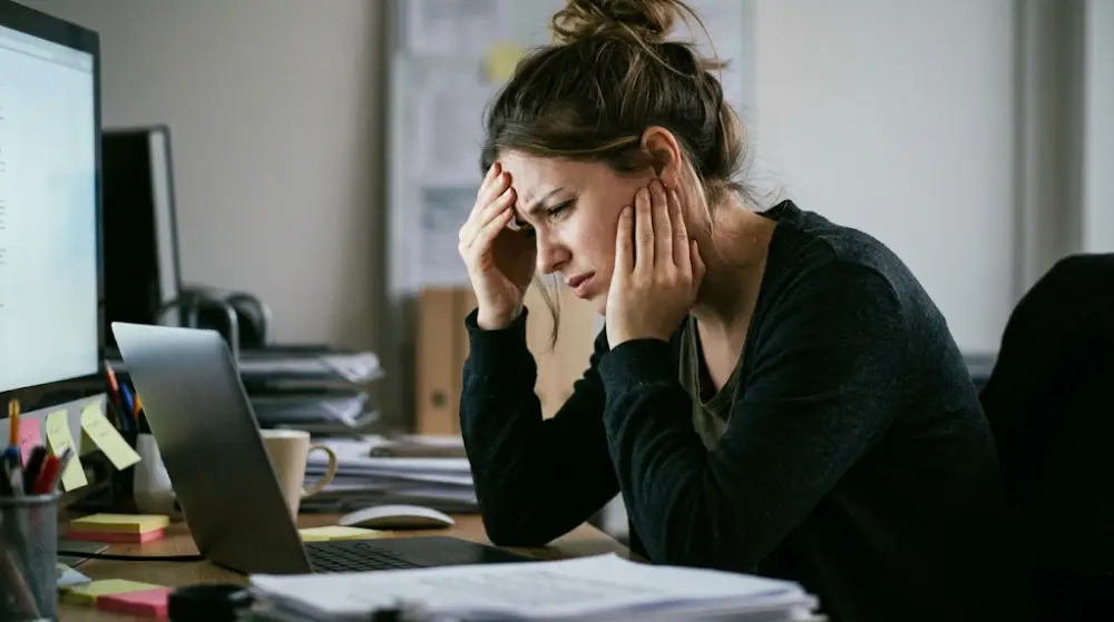 Common Symptoms of TMJ Disorders Woman sitting at a desk rubbing her jaw and temples due to TMJ pain and poor posture.